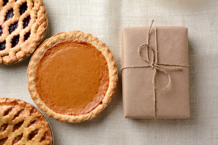 High angle shot of three holiday pies and a wrapped parcel on a burlap tablecloth. The cherry and apple pies run out of the frame with a whole pumpkin pie and package dominate the frame.